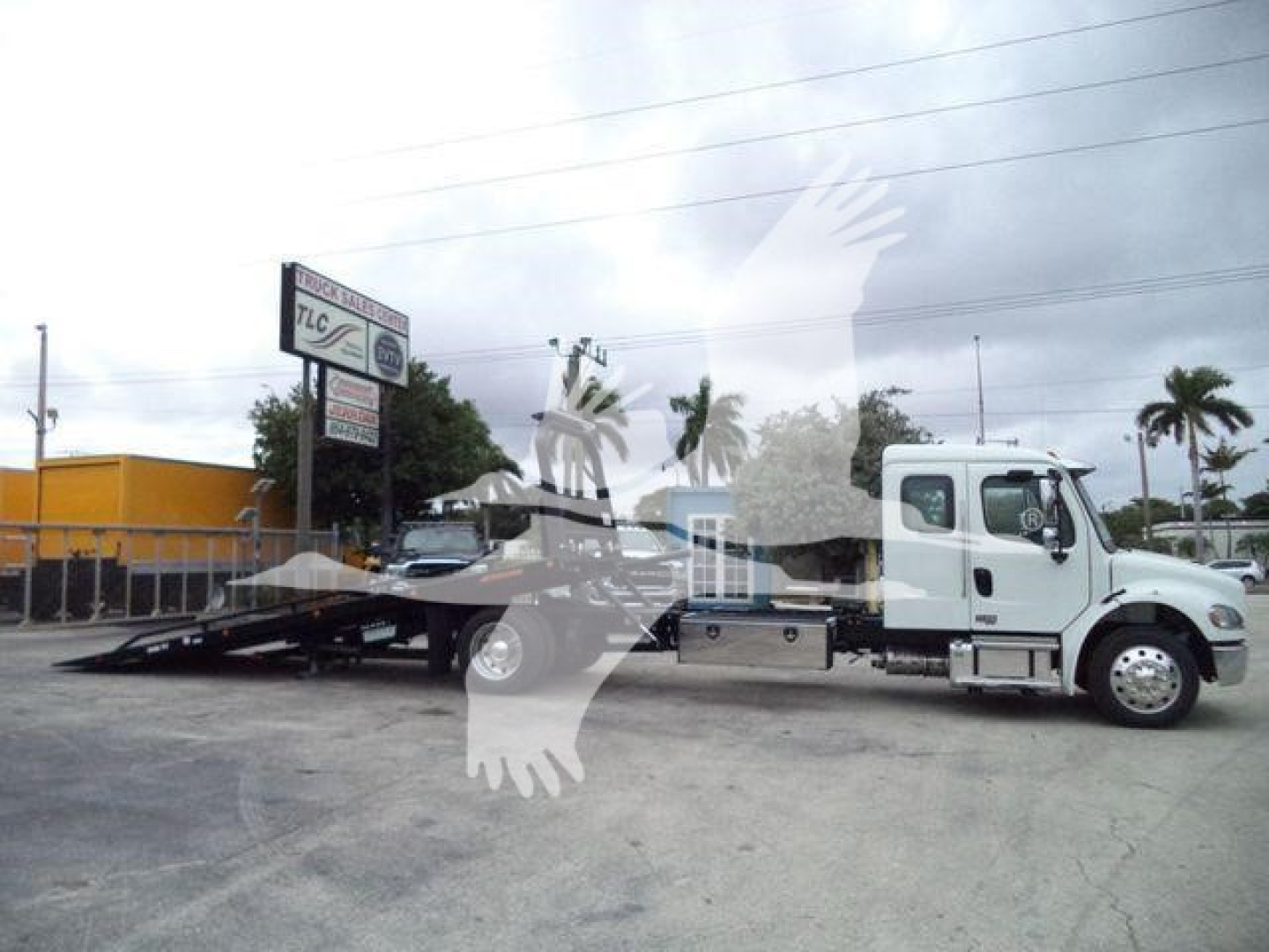 Freightliner M2 106 rollback truck in side view with flatbed and ramp, white cab, light gray equipment; dealership lot in background.