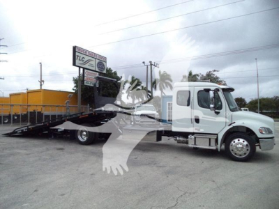 Freightliner M2 106 rollback truck in white with a long flatbed and hydraulic ramps, side view, dealership lot in background.