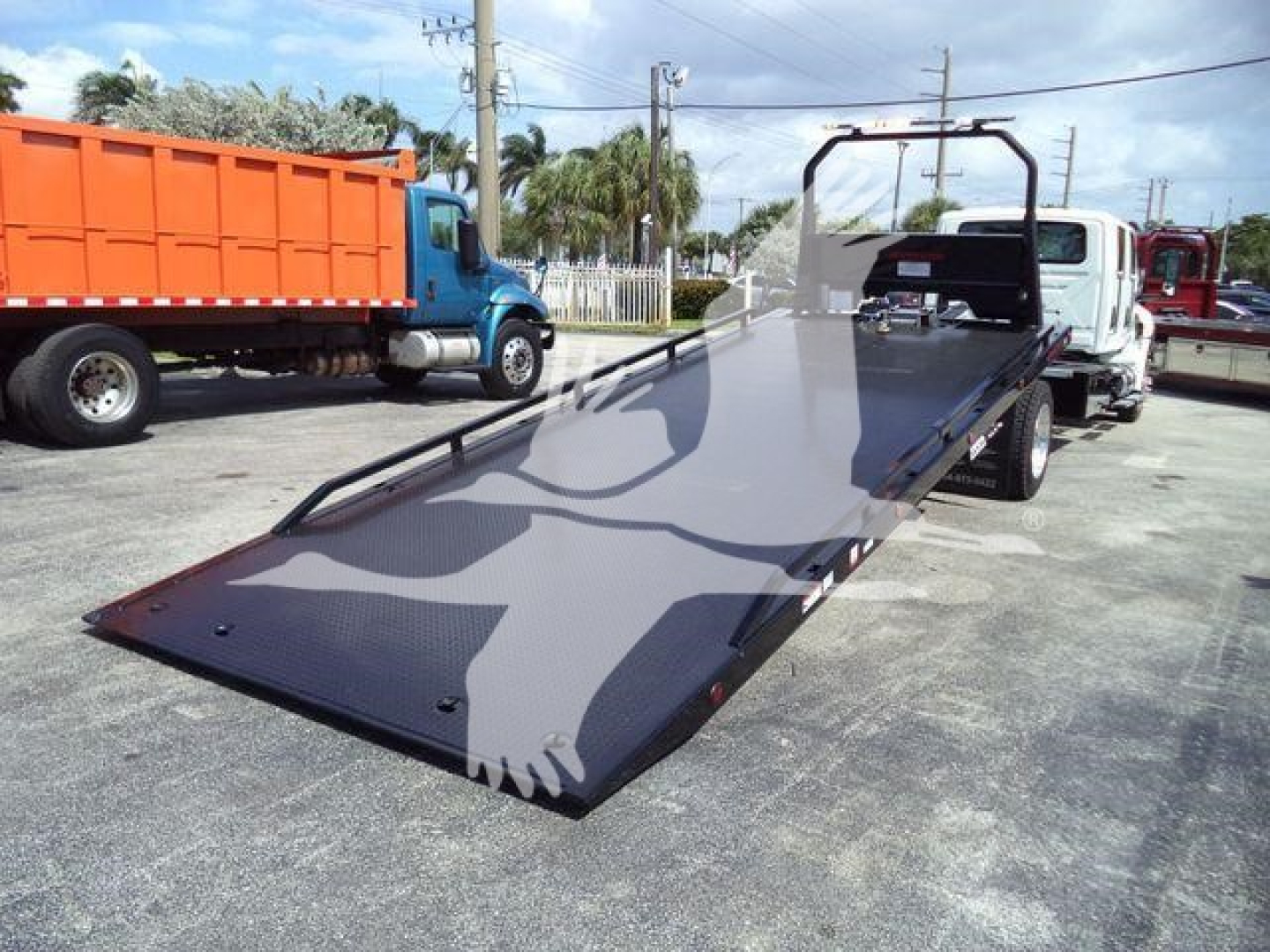 Truck shows a flatbed rollback equipment with a ramp extended from a white cab truck, other trucks in background.