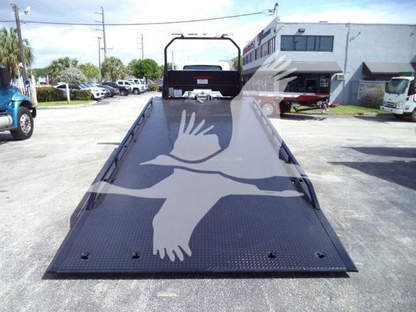 Flatbed tow truck ramp with textured metal surface and a white dove graphic. View shows loading area, rails on sides, rear cab windshield, and surrounding lot with trucks.