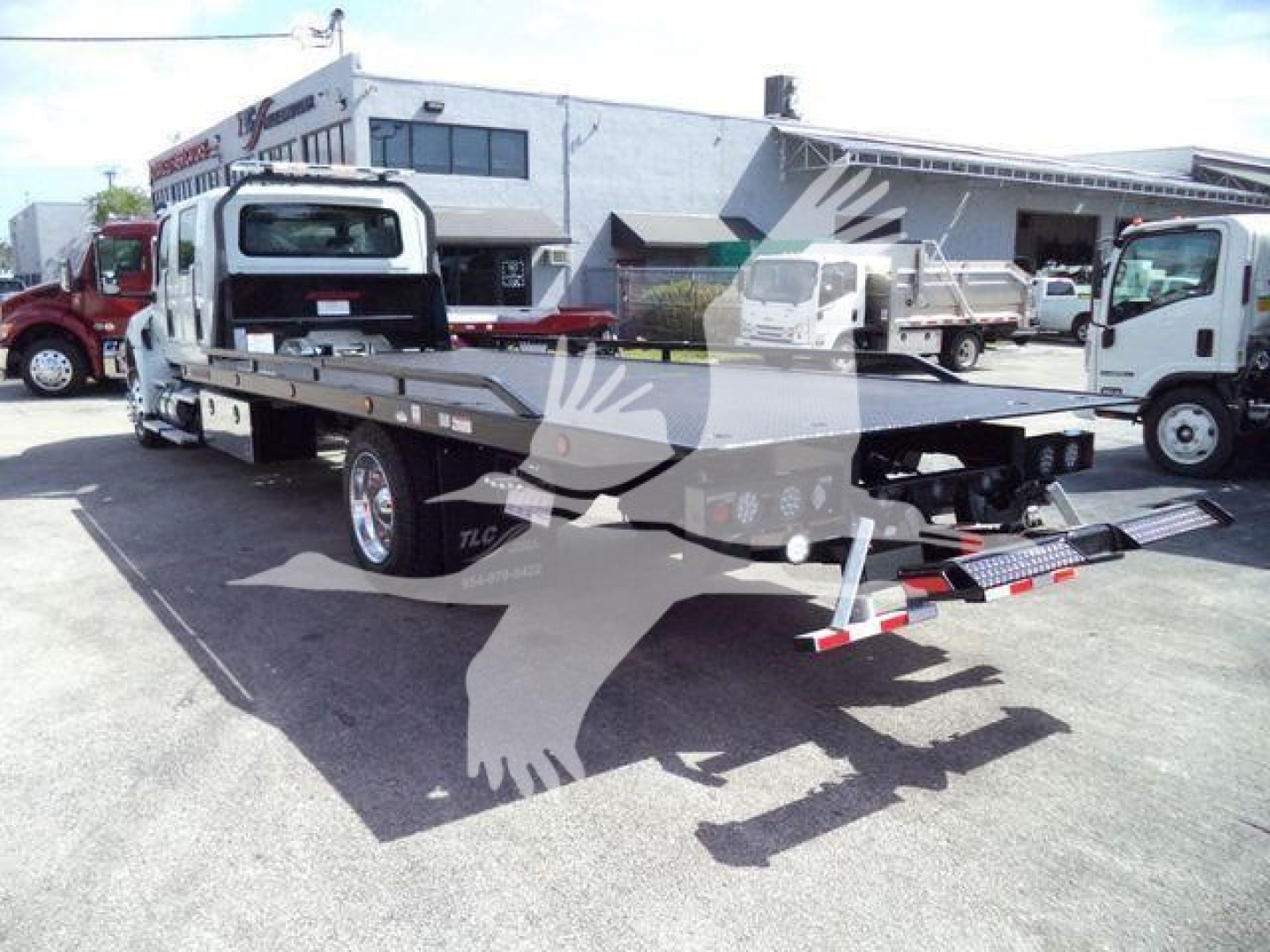 Truck bed with a flatbed tow truck attachment on a white International MV chassis, outdoor lot, other trucks in background.