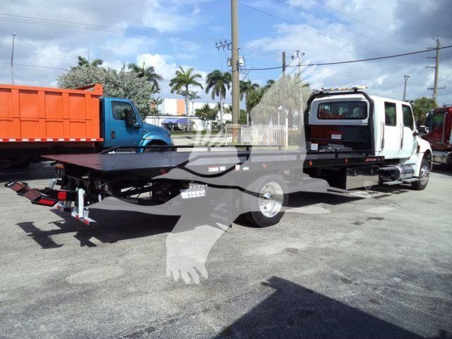 Flatbed tow truck with MV-style truck in background and a white cab; visible text on license plate area and branding not legible.