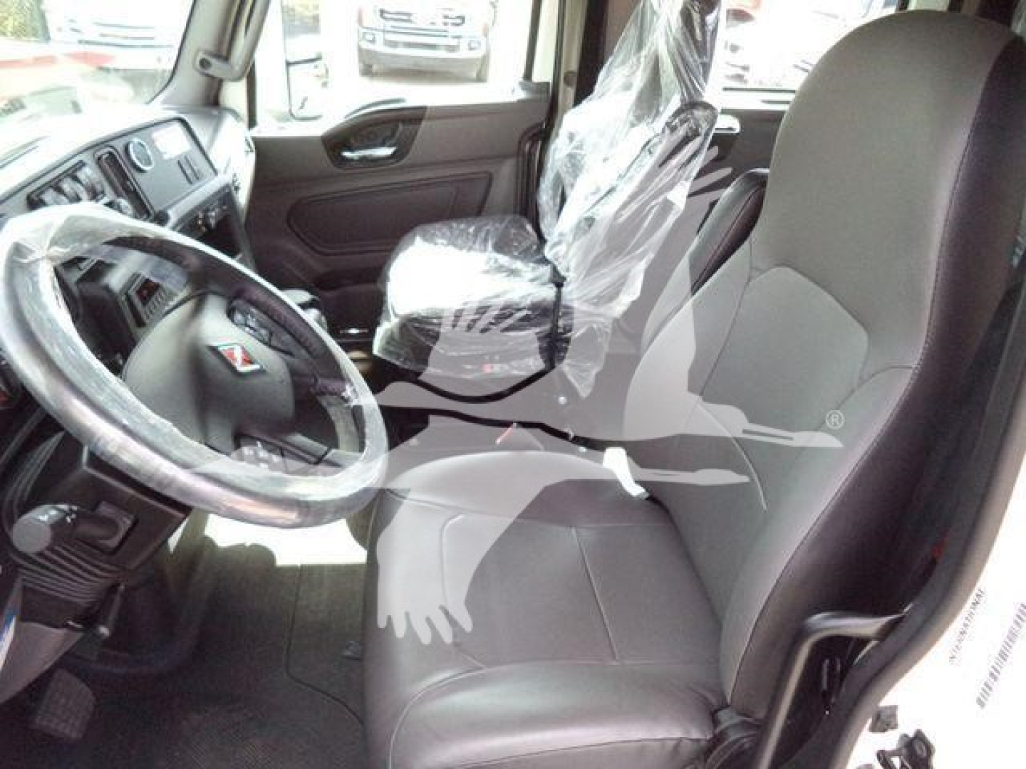 Truck cab interior showing driver's seat with protective plastic, steering wheel, and front passenger seat; plastic-wrapped seat material and dashboard visible.