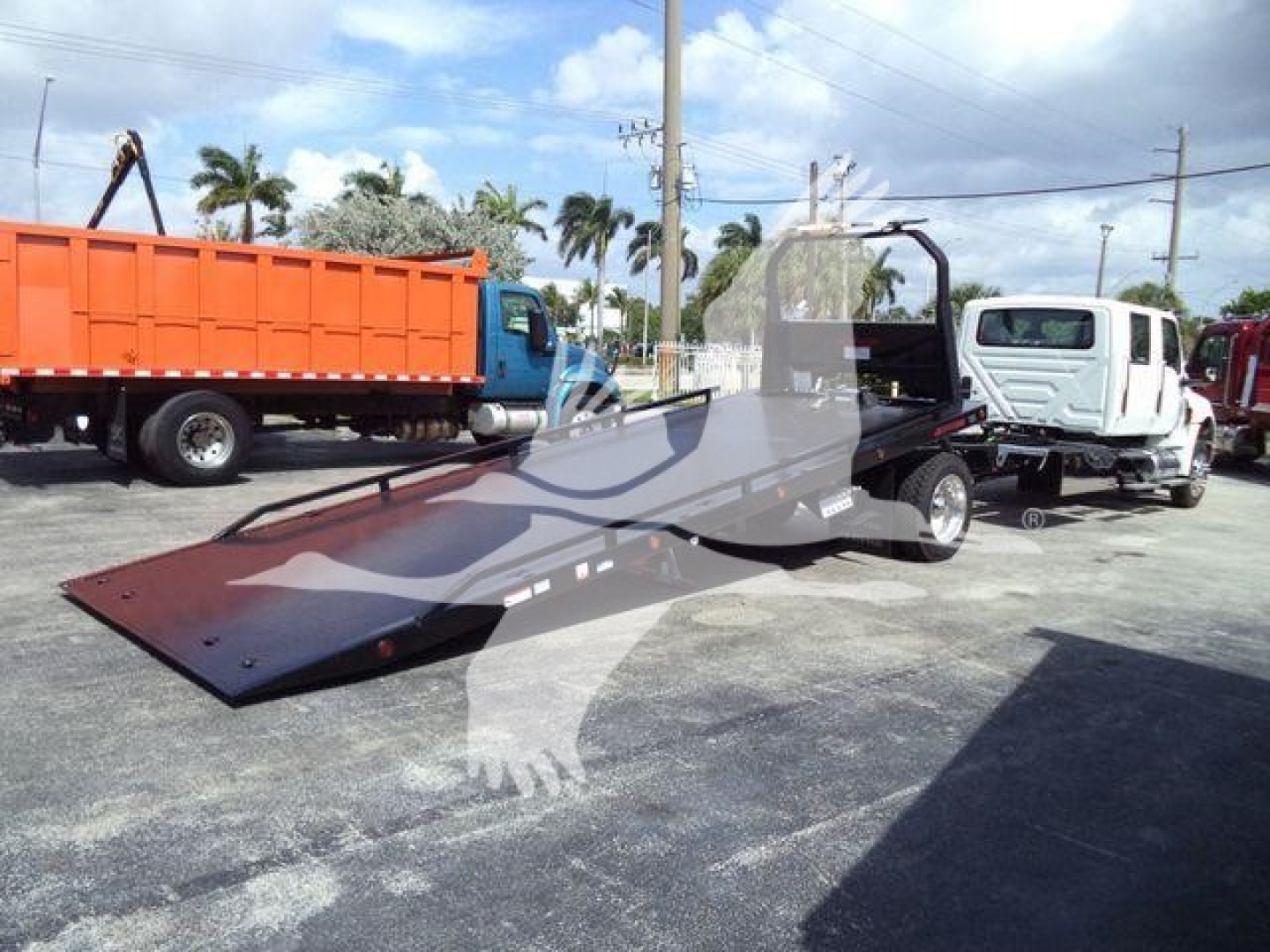 Truck with a ramp/flatbed at a dealership, MV loading equipment, cab in white, large orange dump truck in background