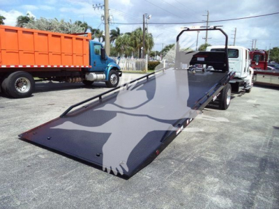 Truck shows a flatbed rollback equipment with a ramp extended from a white cab truck, other trucks in background.