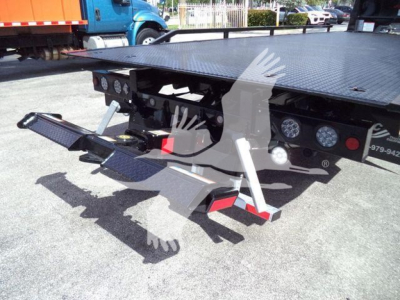 Flatbed rollback truck bed with hydraulic controls and wheel ramps visible, International MV model in the background.