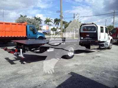 Flatbed tow truck with MV-style truck in background and a white cab; visible text on license plate area and branding not legible.