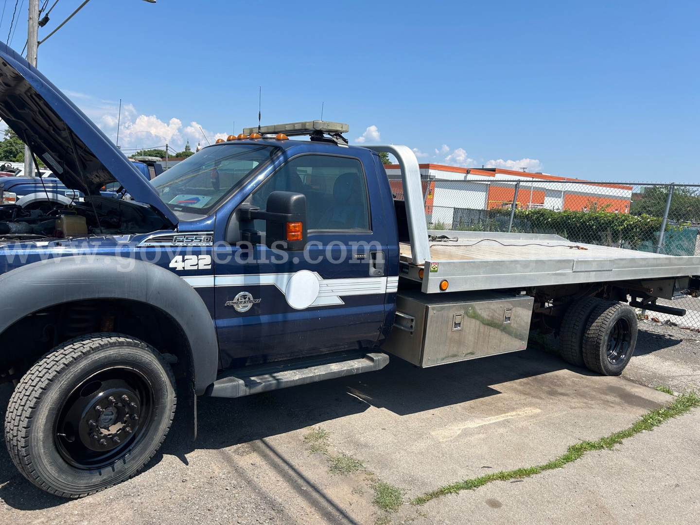 Ford F-550 tow/flatbed truck with hood open and service bed, side decals, and storage box visible.