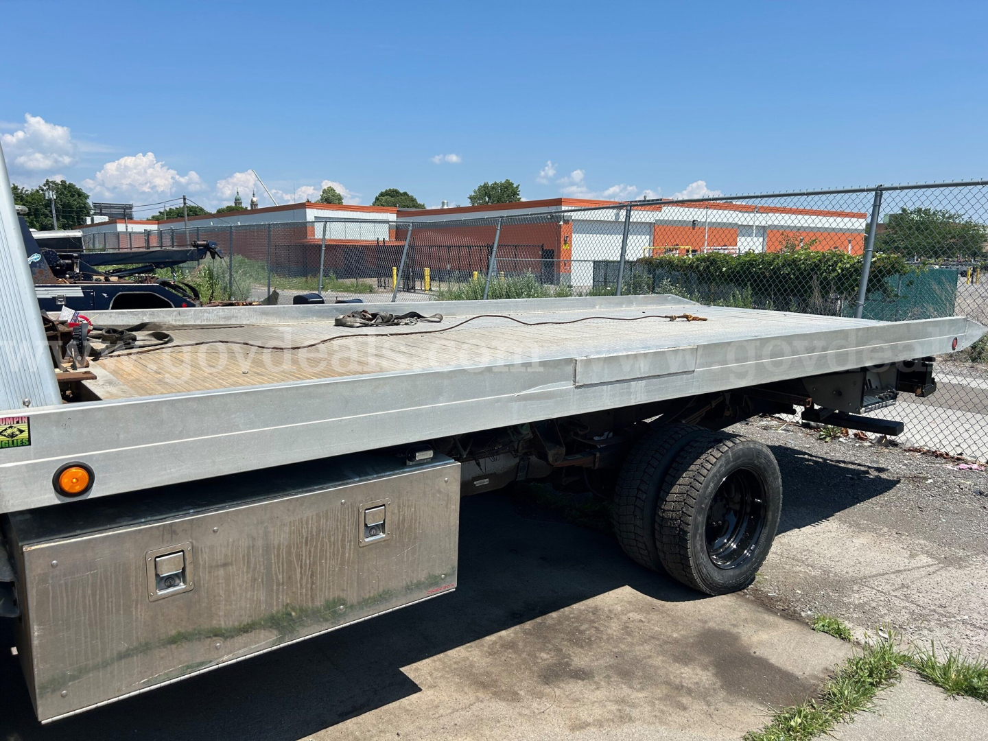 Flatbed tow truck bed with single rear wheel visible, aluminum platform, toolbox on side, chain and hooks on deck, chain-link fence and industrial buildings in background.