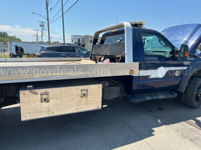 Flatbed tow truck side view with large wooden toolbox under flatbed and text 'www.govdeals.com' on side window; Ford F-550 badge not visible; VIN label not legible.