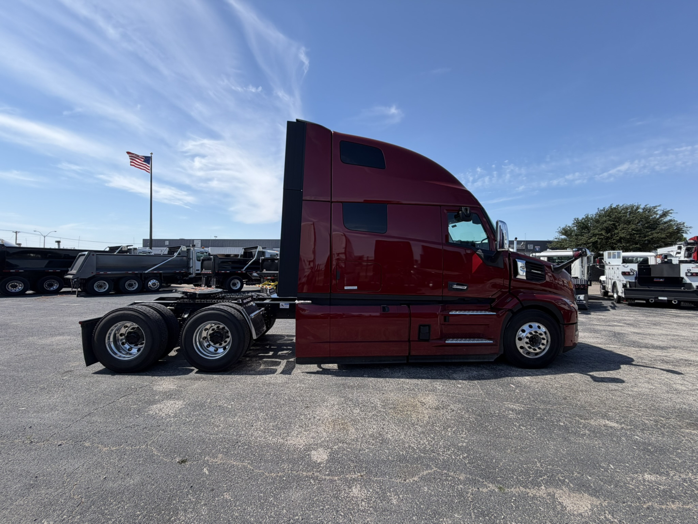 Side view of a maroon 2027 Mack PR64T76H truck tractor with sleeper cab, visible fuel tanks, wheels, and trailing chassis in a dealership lot.
