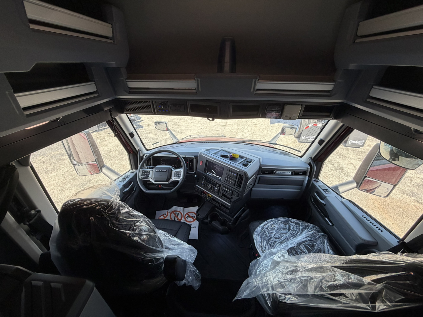 Cab interior of a Mack PR64T76H truck: steering wheel, dashboard, two front seats covered in plastic, various controls visible; overhead storage compartments and windows show outside lot.