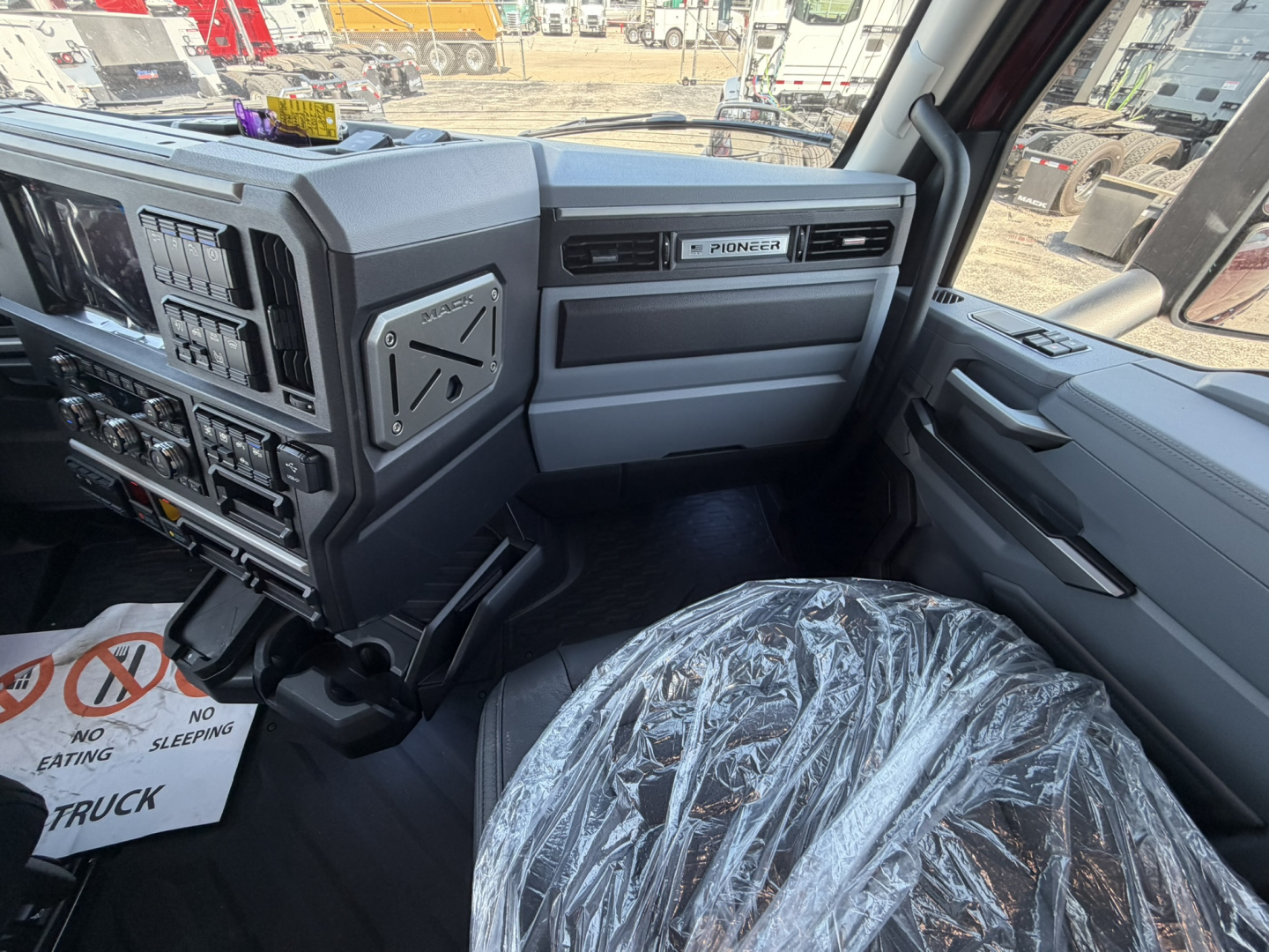 Interior cab view of a 2027 Mack PR64T76H truck showing steering wheel, dashboard with numerous switches, Pioneer infotainment, and plastic-covered driver seat.