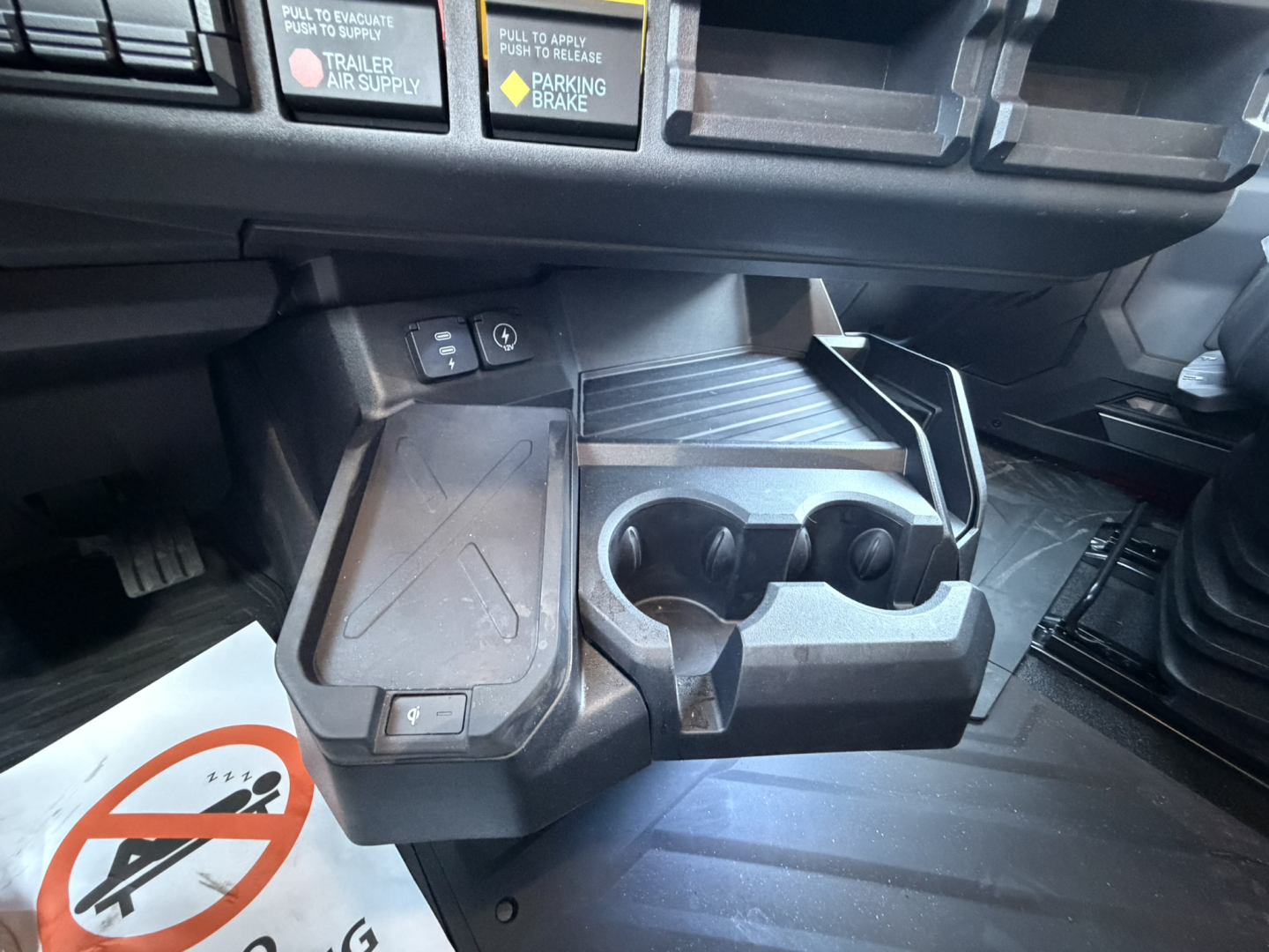 Interior of a Mack truck cab center console with cup holders, storage, and control switches. Visible text includes warning labels and icons.