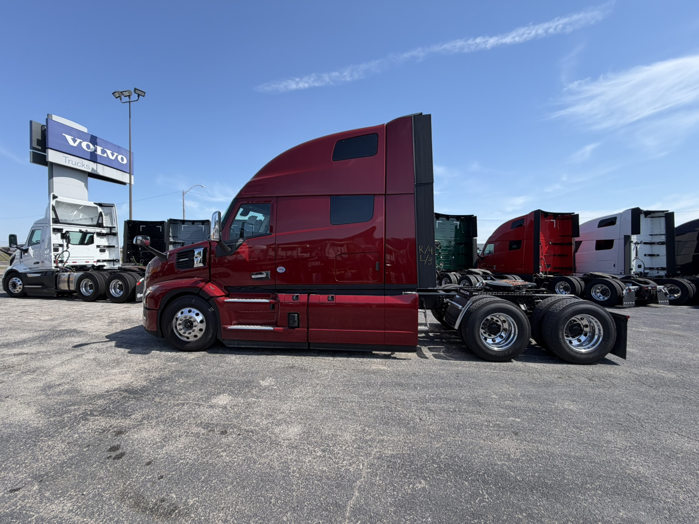 Truck cab side view of a red Mack PR64T76H sleeper tractor, showing long hood, dual rear axles, and chrome wheels, with other trucks in background.