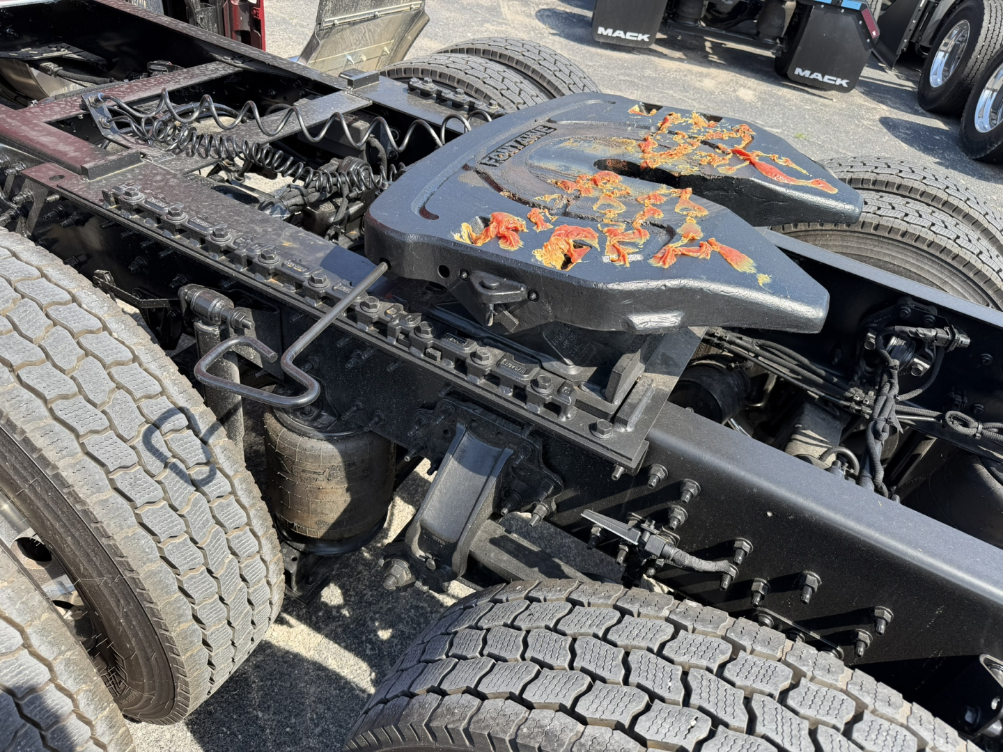 Truck chassis and fifth wheel area of a Mack PR64T76H tractor showing exposed frame, rear wheels, and drivetrain components with some peeling orange paint on a top cover.