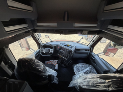 Cab interior of a Mack PR64T76H truck: steering wheel, dashboard, two front seats covered in plastic, various controls visible; overhead storage compartments and windows show outside lot.