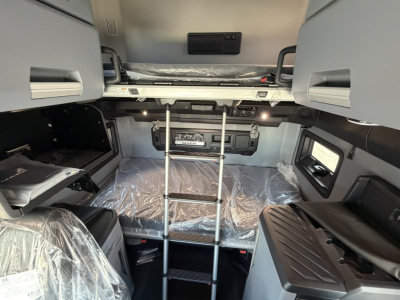 Cab interior of new 2027 Mack PR64T76H sleeper with ladder, bunks, and control panel visible; plastic-covered seating and floor.