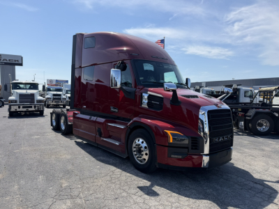Mack PR64T76H conventional sleeper truck in maroon showing cab and front grille with Mack logo and headlights, parking lot with other trucks in background.
