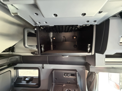 Cab interior of a Mack PR64T76H showing storage compartment with open overhead locker and dashboard controls.