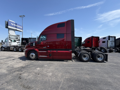 Truck cab side view of a red Mack PR64T76H sleeper tractor, showing long hood, dual rear axles, and chrome wheels, with other trucks in background.