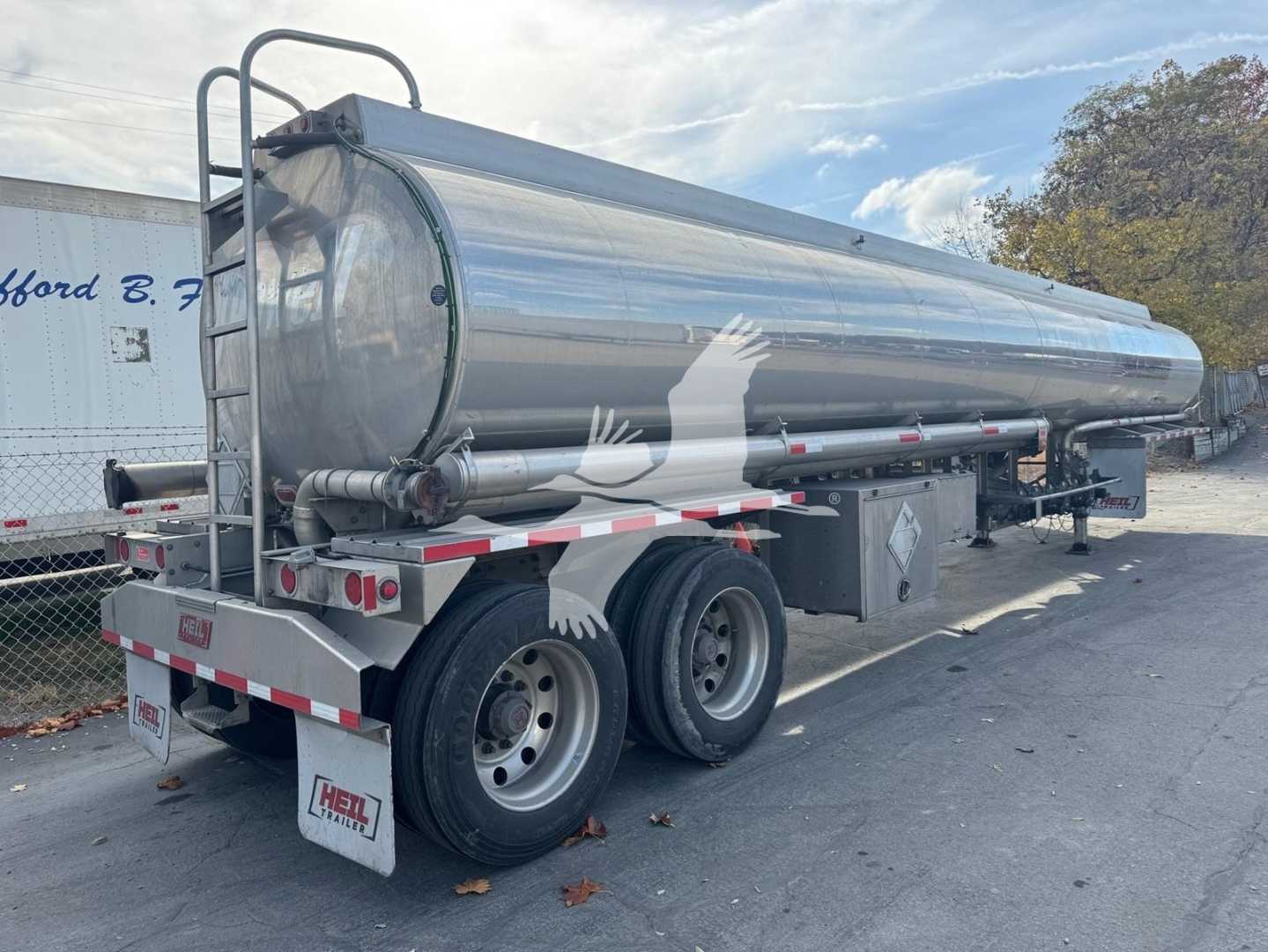 Tank trailer showing cylindrical stainless steel tank with rear access ladder and dual axles. Side text not legible. Off-line pump system under tank.
