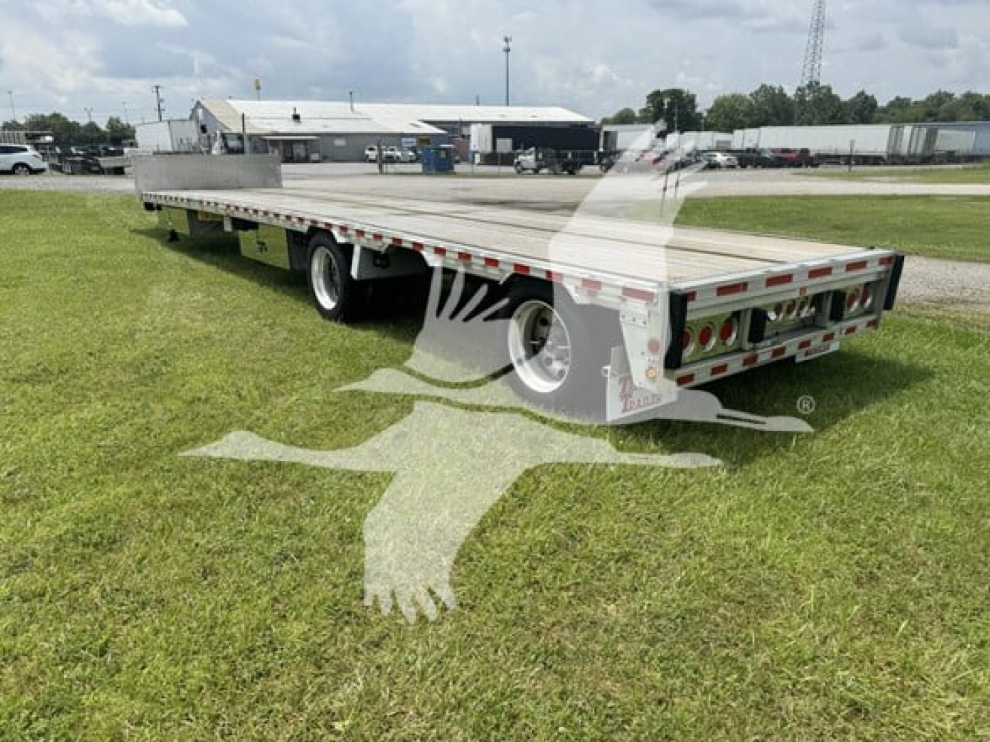 Trailer showing a flatbed with drop deck, rear view showing dual axles and rear end with lights and bumper; image includes grassy lot and industrial buildings.