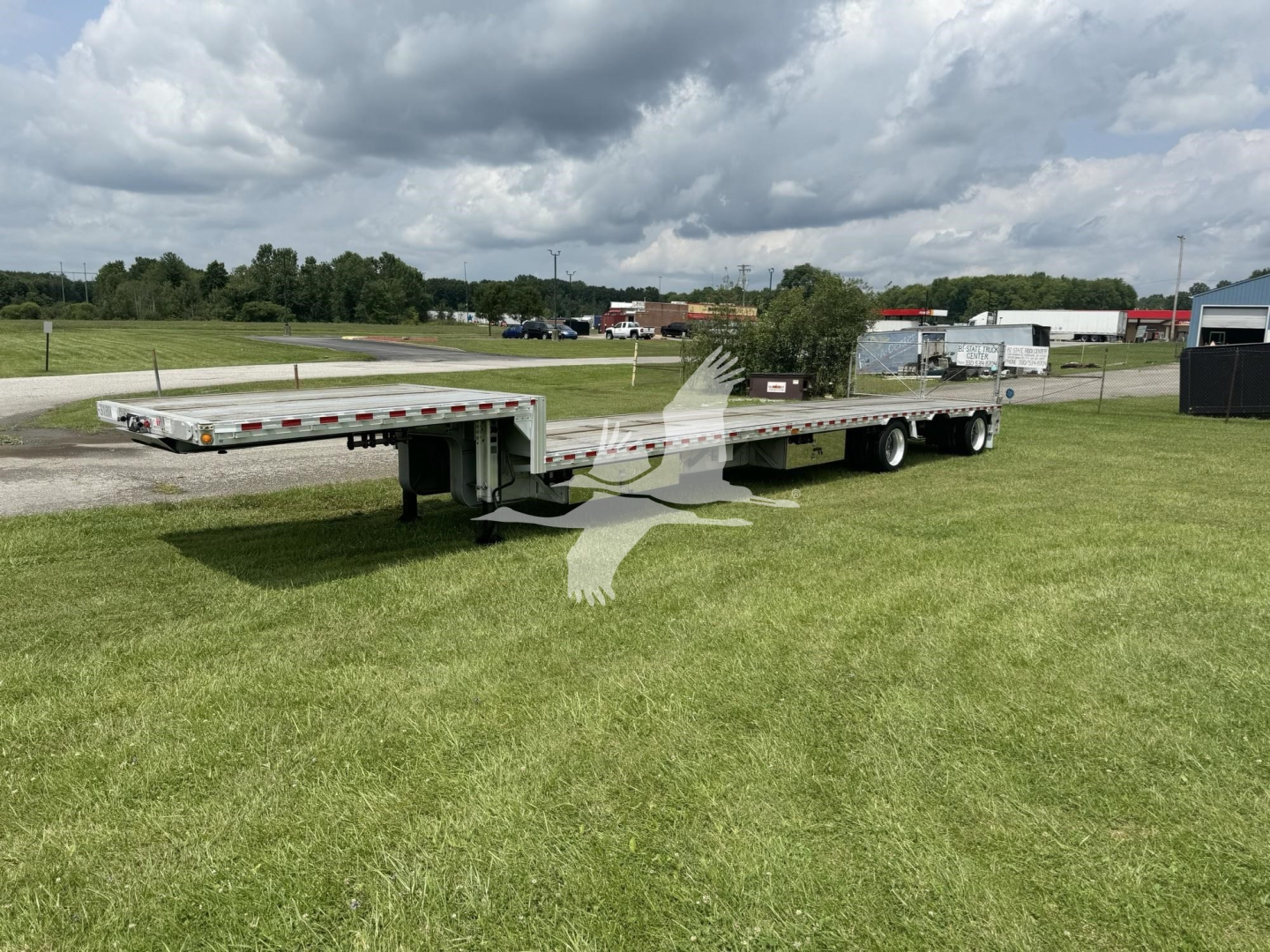 Dropdeck trailer showing long flatbed with dual rear axles and ramps, light gray paint, under a cloudy sky.