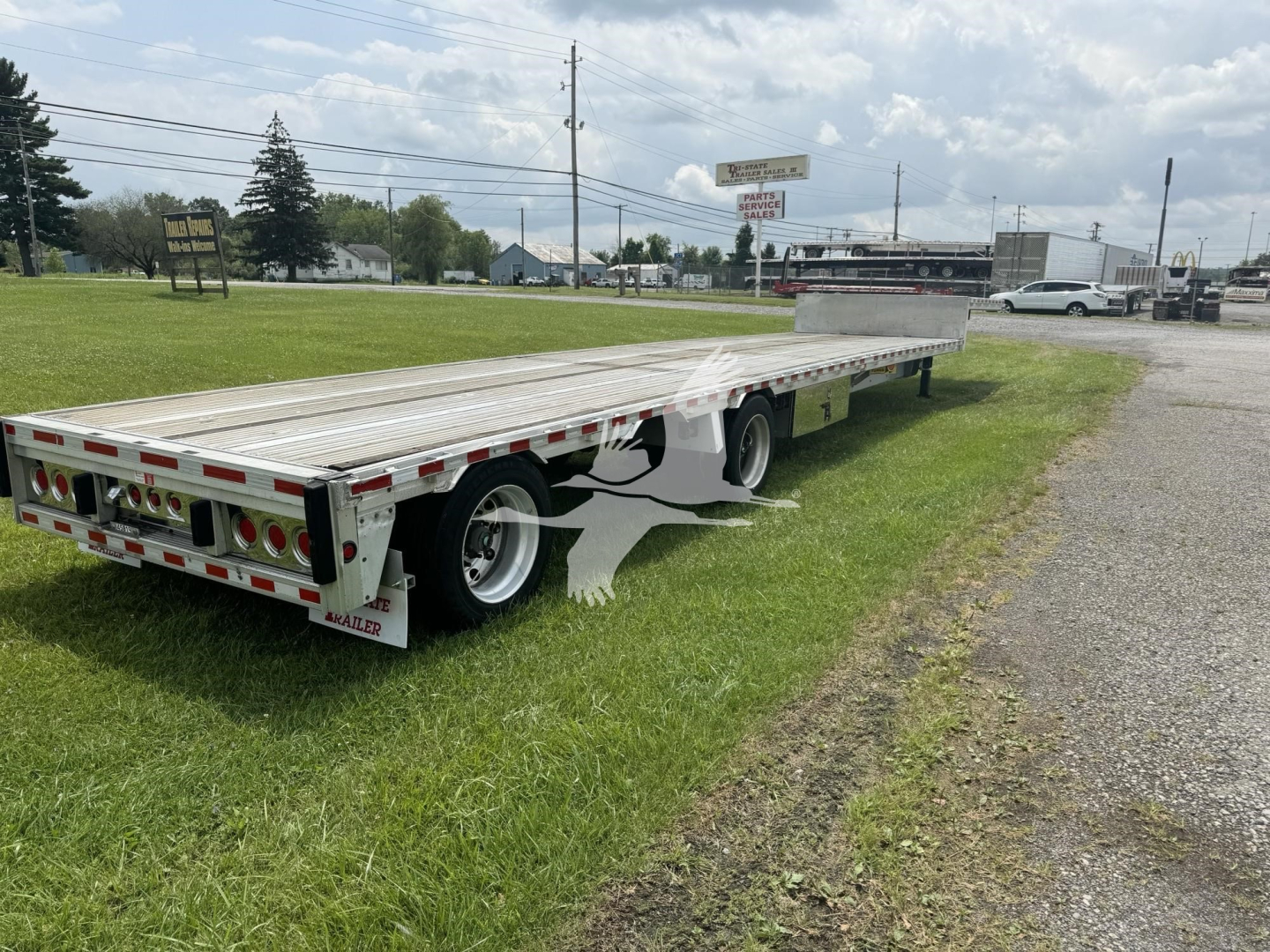 Flatbed trailer in a grassy lot, long aluminum deck with rear lights, reflectors, and a tandem axle. Visible branding on rear bumper and side panels.