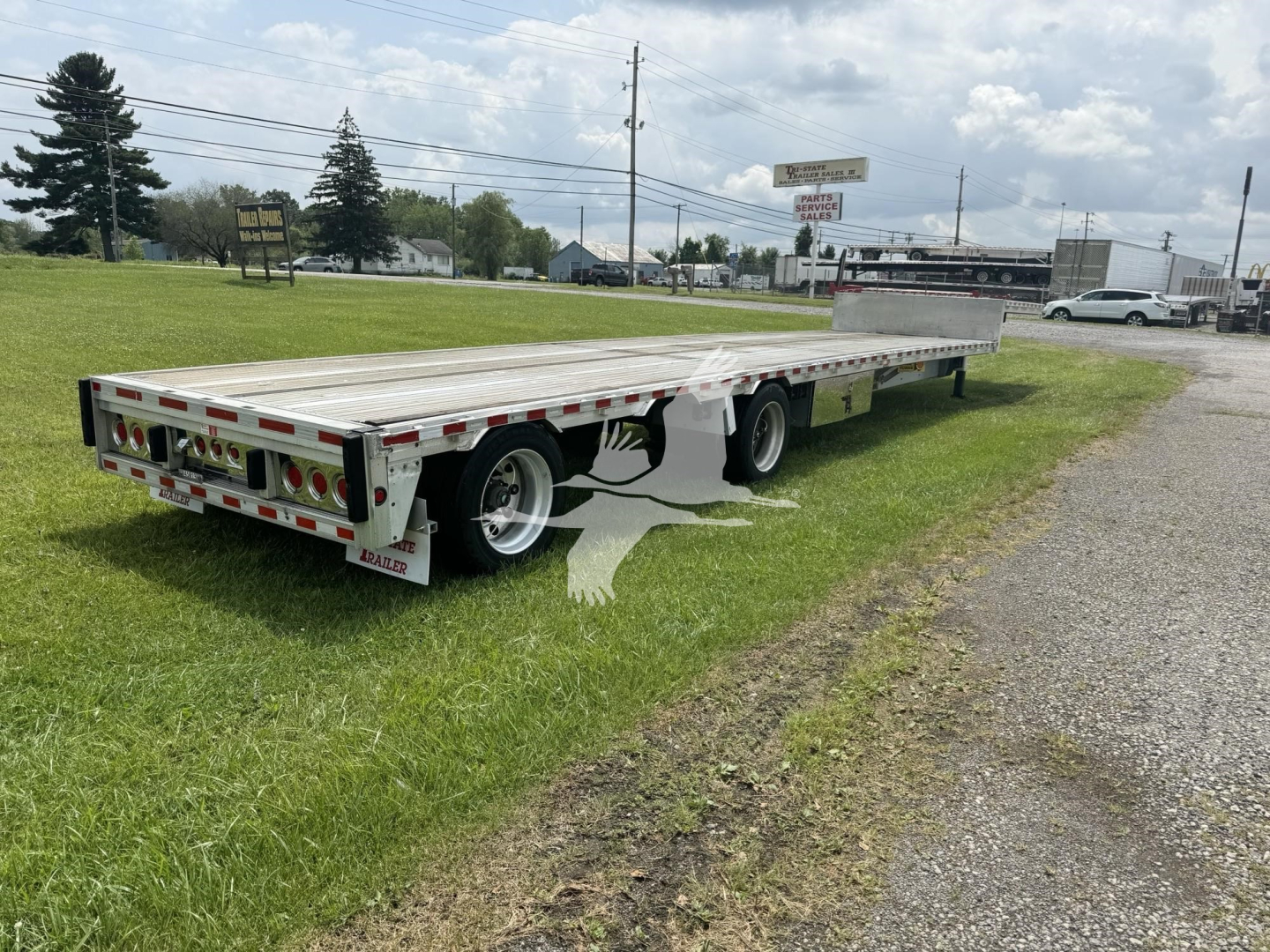 Flatbed trailer with aluminum drop deck, rear lights and dual axles. Visible text on bumper and side panels includes dealer stickers and reflective tape.