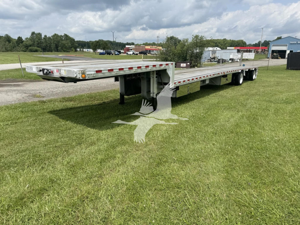 Flatbed trailer with drop deck, side rails, and tandem axles in an open lot. Visible markings include reflectors and lights along the side. No legible brand text or numbers clearly readable on the trailer.