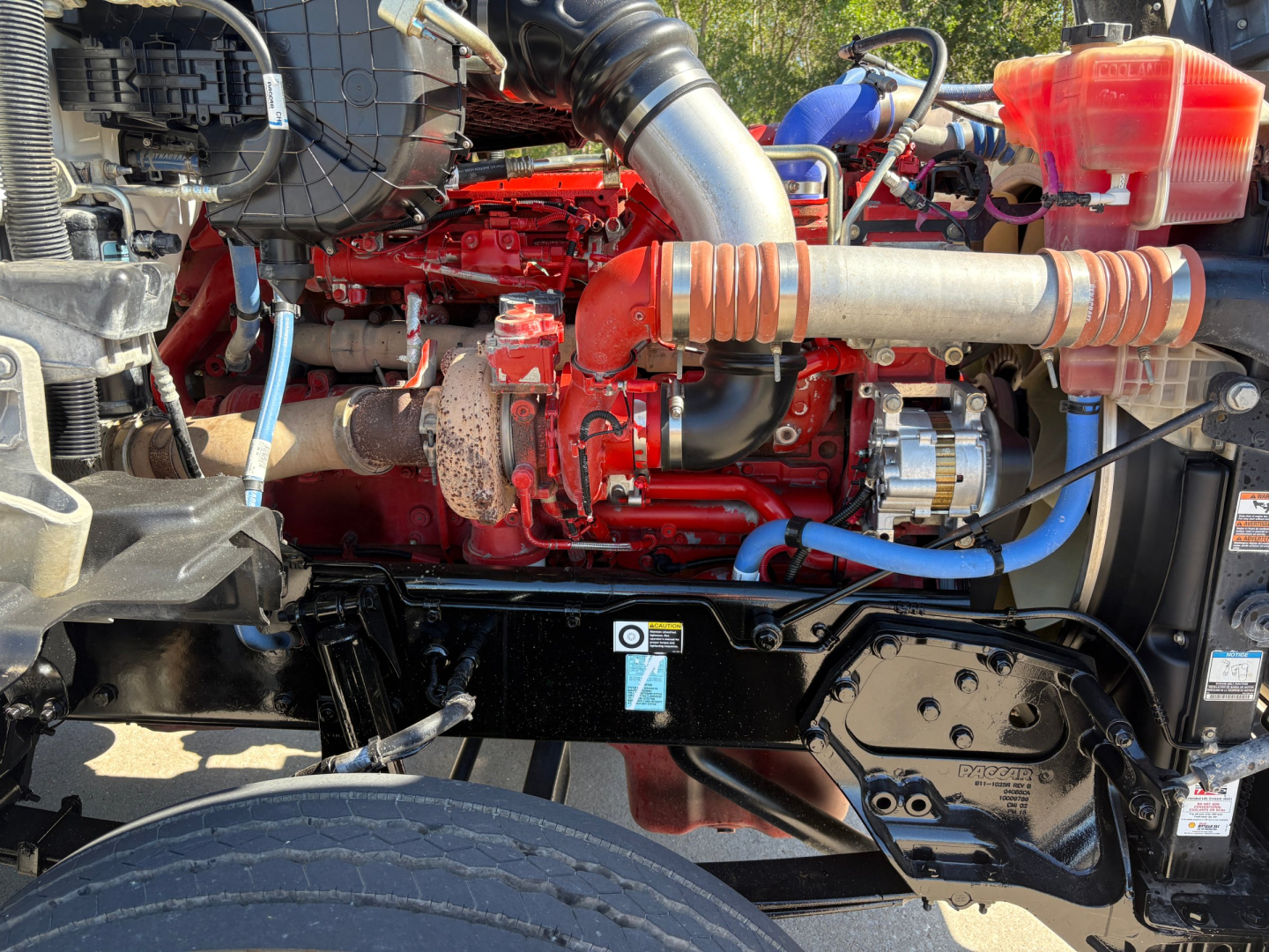 Kenworth T880 engine bay showing red engine, black chassis, exhaust manifold, hoses, battery and coolant reservoir cap.