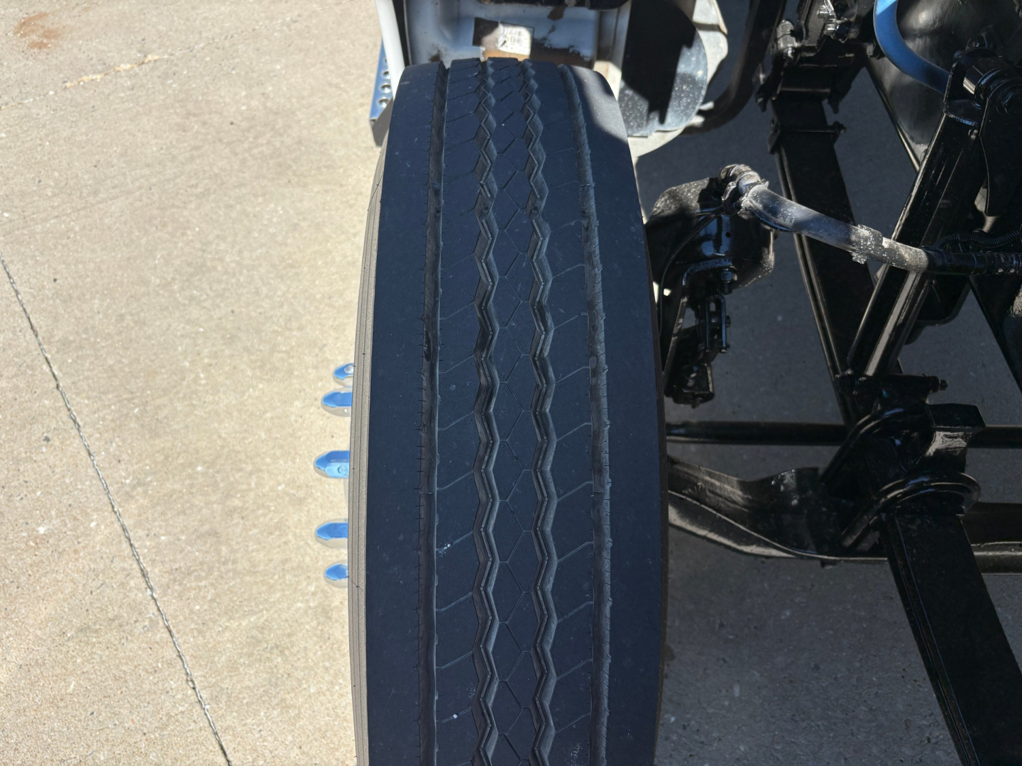 Truck tire view with side of chassis and suspension components. The tire shows a tread pattern and blue fasteners visible along the rim area.