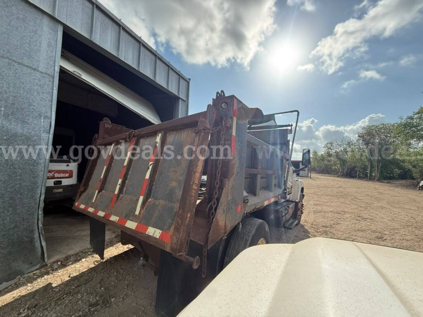 Dump truck bed of a GMC dump truck with a rusted steel bed and hydraulic lift, parked near a warehouse. Text on image includes a watermark 'govdeals.com' partially visible.