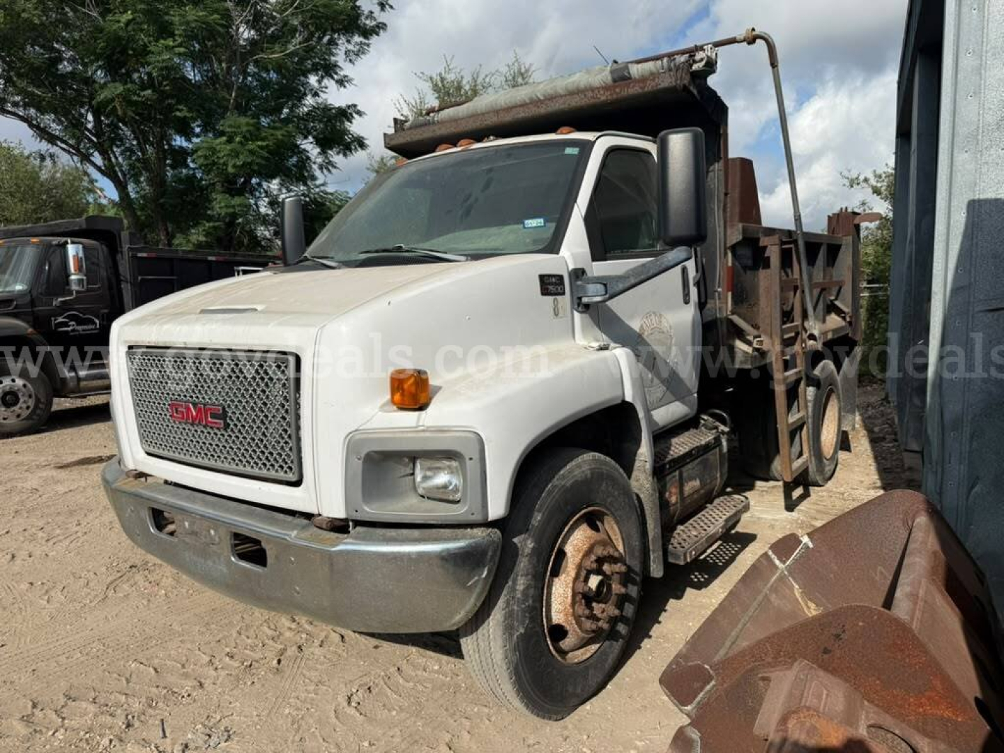 GMC truck in yard with dump bed, white cab, front grille GMC emblem, visible rust on rear wheel area and dump bed; safety mirrors on sides.