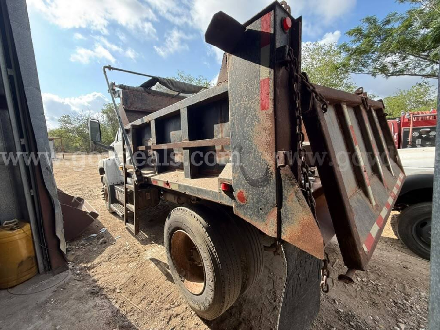 Dump truck bed side view showing GMC dump truck with hydraulic bed and chain hooks, rear tailgate, mud and dirt, sky and trees in background.