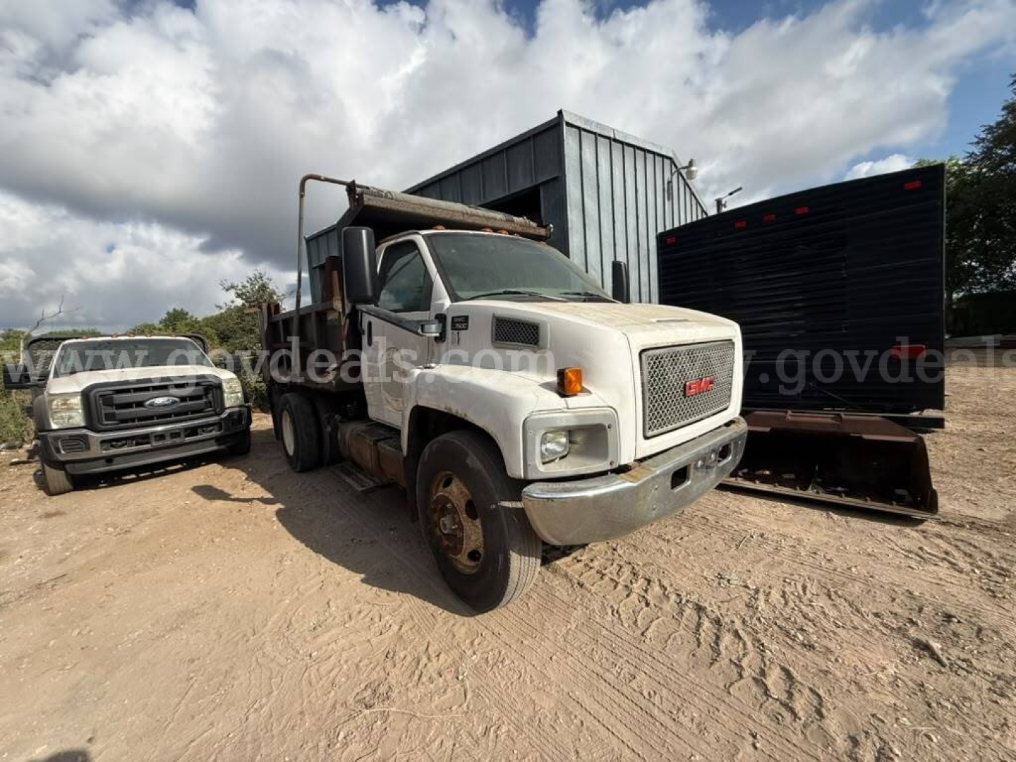 GMC dump truck in yard showing front grille, white cab, and dump bed with rust; gravel ground and other vehicles in background.