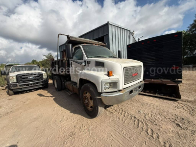 GMC dump truck in yard showing front grille, white cab, and dump bed with rust; gravel ground and other vehicles in background.