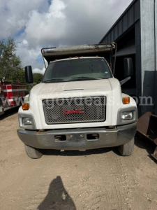 GMC dump truck front view showing grille with red GMC emblem, rectangular headlights, and bumper; white cab with dump body partially visible.