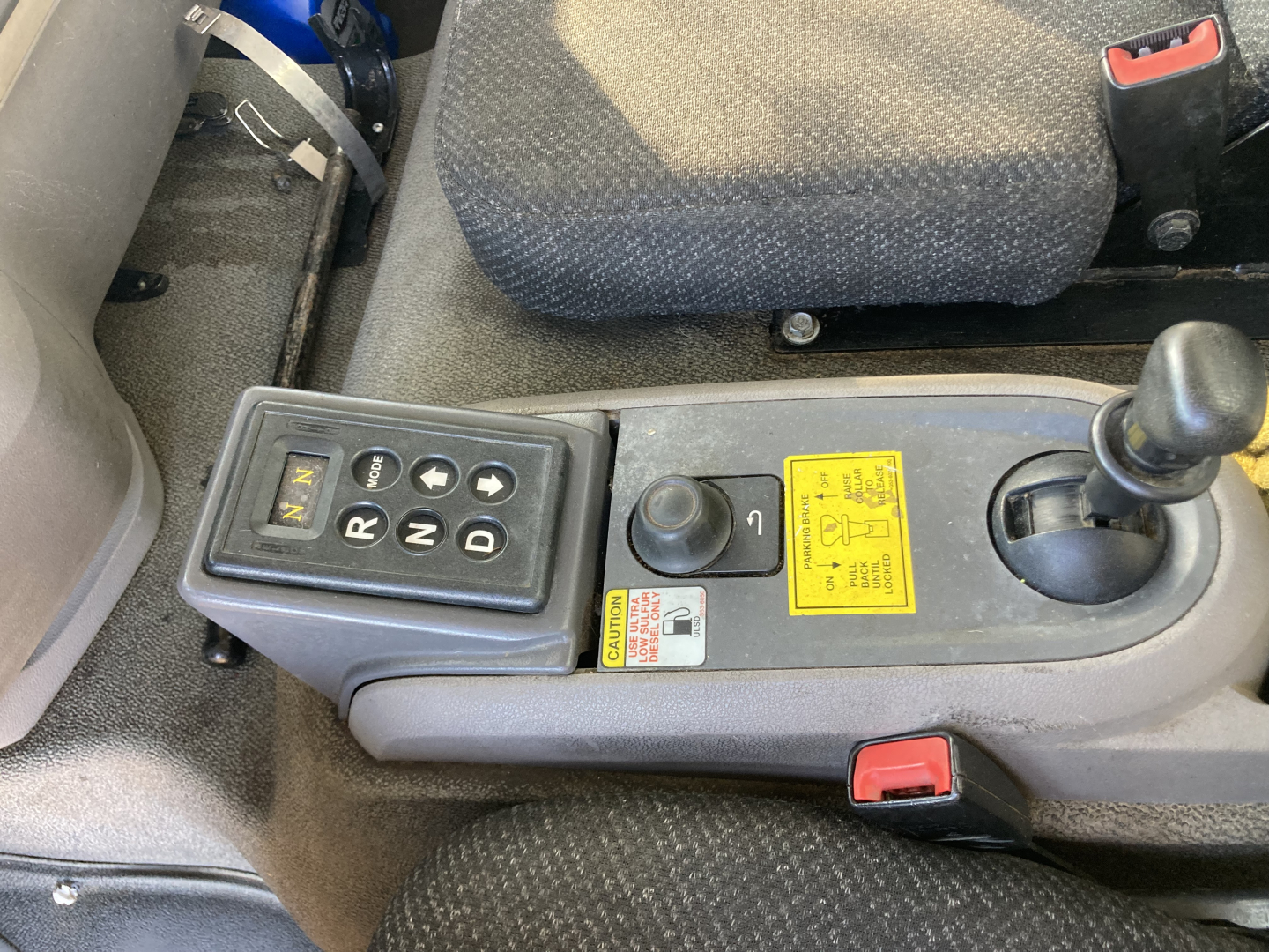 Peterbilt cab interior showing gear selector with multiple modes, joystick control, yellow caution label, red seat belt buckle, and black shift lever in center console.