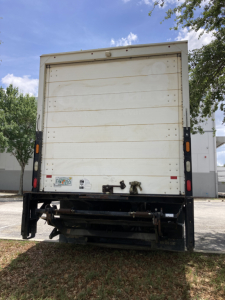Rear view of a white Peterbilt box truck with license plate area, rear bumper, and taillights. Visible text includes small stickers on lower left of the cargo door.