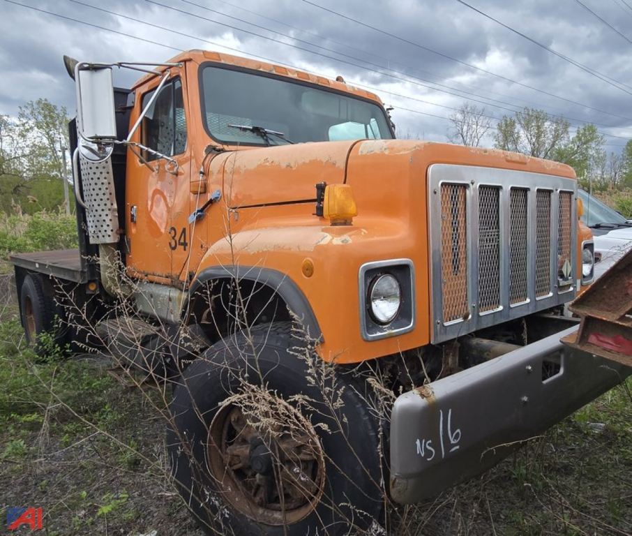 1997 International 2574 dump truck flatbed in orange with large front grille and bumper; visible side step, front left wheel, and overgrown vegetation around vehicle.