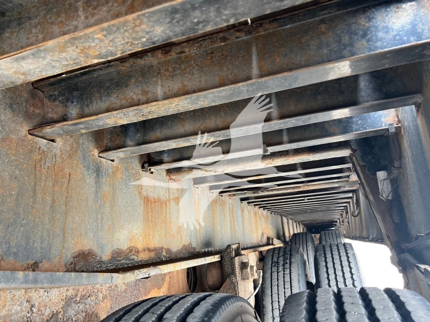 Trailer undercarriage with dual axles and wheels, rusty metal beams, visible tires, and rectangular frame structure of a Landoll trailer.