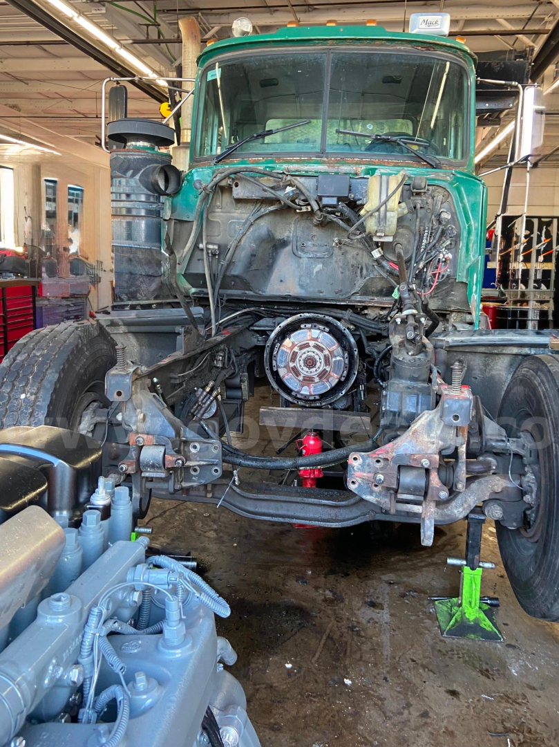 Mack RD688S truck front view with engine bay open; exposed drivetrain, hydraulic components, and wiring visible; green cab and workshop environment.