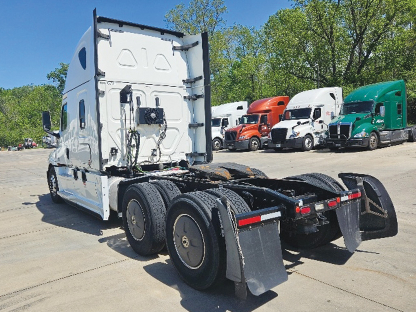 Truck tractor rear view showing fifth wheel and dual rear axles with mud flaps and white cab, Freightliner Cascadia sleeper tractor in background.