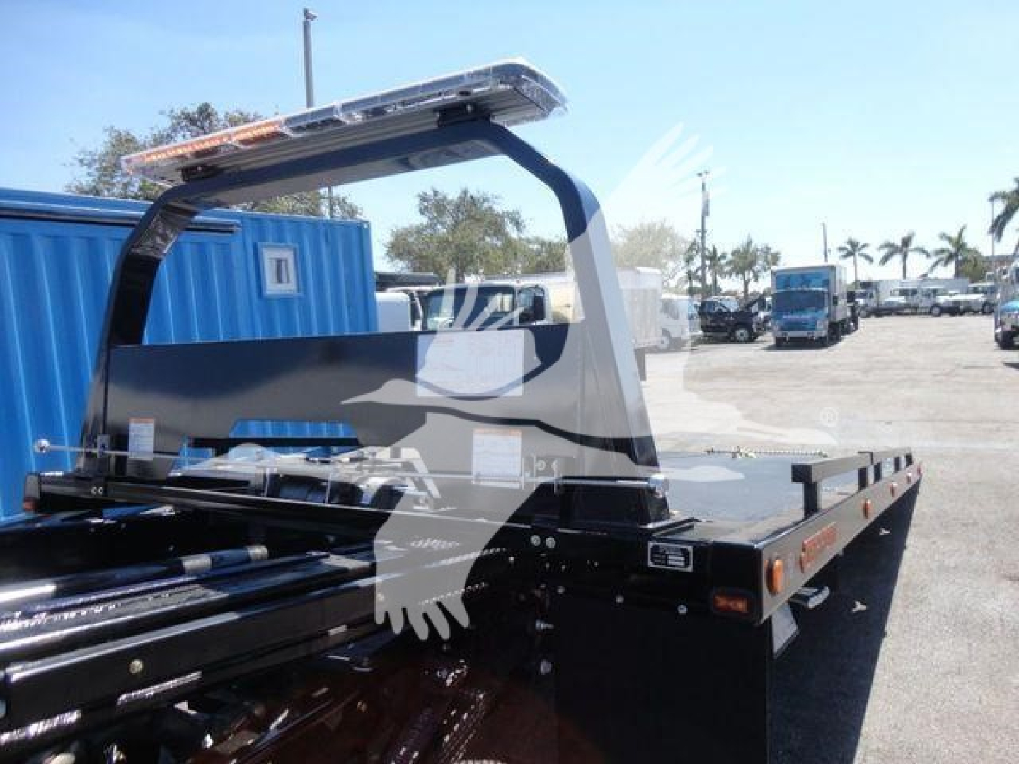 Ram 5500 rollback flatbed truck with cab-over rear setup, black flatbed, light bar, and rear reflectors. Visible stickers and safety rails along bed edges.