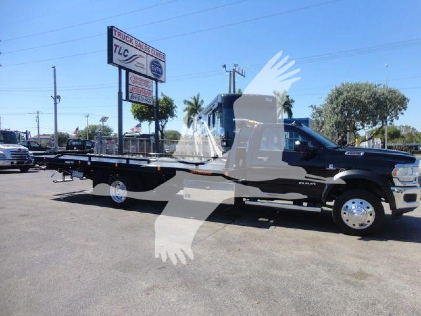 Ram 5500 rolling tow truck on lot with rollback bed, black cab, white wheels, dealership sign in background. Visible text on signs includes TLc and truck sales but small.