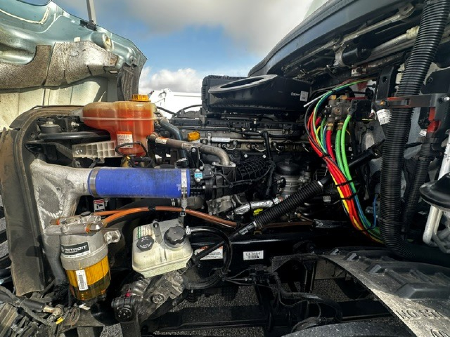 Freightliner Cascadia engine bay showing left-side components including orange coolant reservoir, blue silicone hose, multiple colored electrical wires, fuel filter, and compressor; view from driver side with open hood.
