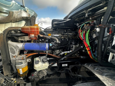  Freightliner Cascadia engine bay showing left-side components including orange coolant reservoir, blue silicone hose, multiple colored electrical wires, fuel filter, and compressor; view from driver side with open hood.