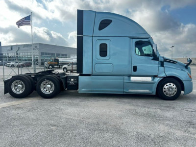 Truck side view of a blue 2023 Freightliner Cascadia 126 sleeper cab tractor with tandem rear axles in a dealership lot.
